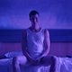 A man wearing an undershirt sits at the edge of a bed in a blue-tinged room.